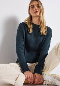 Dunkelblauer Strickpullover mit gerippten Bündchen, kombiniert mit weiten beigen Hosen und weißen Sneakers. Das Model sitzt mit übereinandergeschlagenen Beinen.