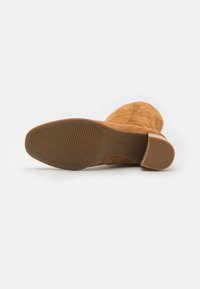 Botte cuissarde en suède couleur beige, avec un bout rond, une semelle plate et une semelle extérieure en caoutchouc texturé. Elle présente un talon bloc lisse pour plus de stabilité.