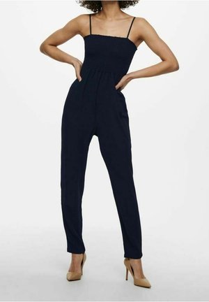 Granatowy kombinezon z cienkimi ramiączkami, marszczonym gorsetem i zwężanymi nogawkami; noszony z cielistymi szpilkami. Gładka tkanina i dopasowany fason.