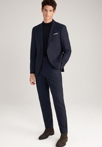 Marineblauer Anzug mit taillierter Passform, ausgestattet mit einem Revers, Ein-Knopf-Verschluss und einer Brusttasche. Kombiniert mit einem schwarzen Rollkragenpullover und braunen Stiefeln.