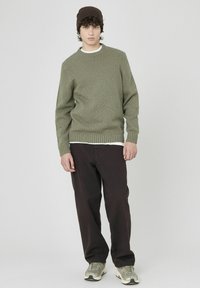 Pull en laine olive avec un col rond, des poignets et un ourlet côtelés, associé à un pantalon ample marron foncé et des baskets grises.