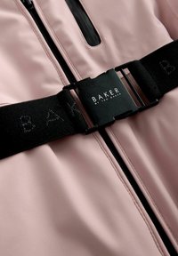 Gaiši rozā jaka ar spīdīgu apdari, kurai ir melna rāvējslēdzēja un jostas detaļas. Jostas plastikāta sprādze ir ar uzrakstu "BAKER", kas drukāts baltā krāsā.