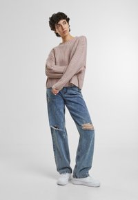 Hellrosa, gerippter Strickpullover kombiniert mit hellblauen, verwaschenen Jeans aus Denim mit großen Rissen. Weiße Sneakers ergänzen das Outfit.