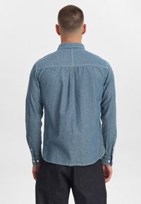 Camicia in denim blu con maniche lunghe, colletto a punte e dettagli con pieghe sul retro. Presenta bottoni bianchi sui polsini e un orlo curvo.