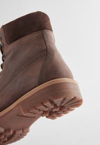 Botte en cuir marron avec col en daim, design à lacets et semelle en caoutchouc robuste avec des motifs de traction profonds. Détails cousus le long du côté.