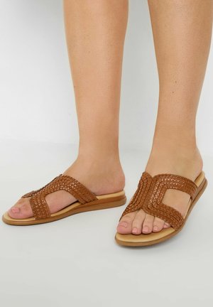 Brown woven slide sandals featuring a flat sole, open toe design, and cross-strap detailing. The straps have a textured finish for grip.