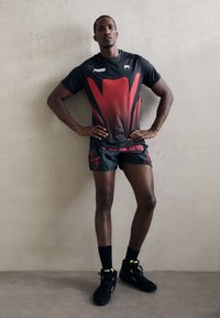 Sportoutfit mit einem schwarzen kurzärmligen Hemd, das ein rotes Farbverlauf- und Punktmuster aufweist, kombiniert mit schwarzen Shorts und schwarzen Schuhen mit neonfarbenen Akzenten.
