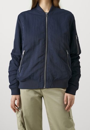 Femme portant une veste bomber bleu marine avec fermeture éclair et un pantalon cargo kaki avec une poche zippée sur la cuisse.