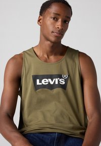 Olive žalios spalvos marškinėliai be rankovių su juodu Levi's logotipu kontrastingoje formoje priekyje; glotni tekstūra.