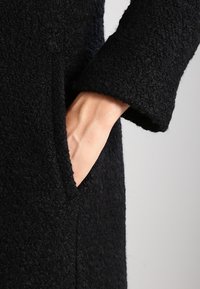 Mano inserita nella tasca di un cappotto nero testurizzato, che mette in mostra il dettaglio della manica e del tessuto.
