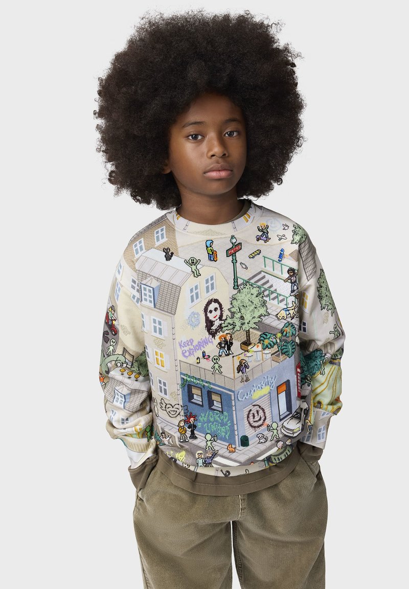 Enfant avec une grande afro portant un sweat coloré illustré avec des bâtiments de la ville, des personnes, des arbres et des scènes de rue, debout les mains dans les poches.