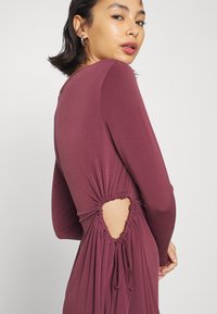 Vestido longo de manga comprida na cor malaquita, com detalhes franzidos e um recorte na cintura. Tecido suave e elástico com bordas ajustadas e um design fluido.