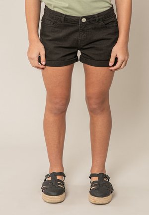 Personne portant un short noir retroussé et des sandales à plateforme noires à lanières, debout devant un fond beige uni.