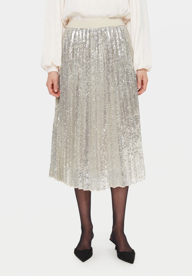 Sömlös midi kjol med silverfärgade paljetter och plissé, med en elastisk midja, i kombination med svarta strumpbyxor och spetsiga svarta skor. Har en glänsande, reflekterande yta.