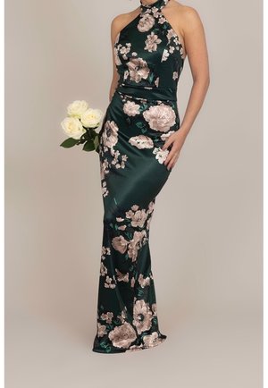 Bezrekawna sukienka w odcieniu ciemnej zieleni w kwiaty, z wiązaniem na szyi, noszona przez osobę trzymającą trzy białe róże na jednolitym tle.