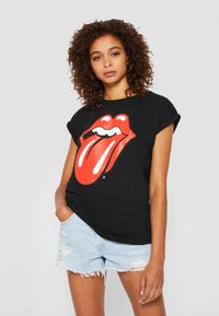 Sort T-shirt med et stort rødt Rolling Stones-logo med hvide detaljer. Sat sammen med lyseblå slidte denimshorts.