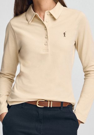 Polo beige à manches longues avec boutons et petit logo brodé, associé à un pantalon foncé et une ceinture marron avec boucle dorée.