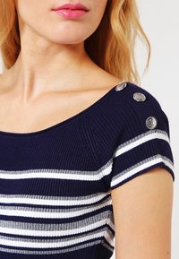Marinblå och vit randig stickad topp med korta ärmar, prydd med metallknappar på axeln som accent. Slät textur och figurnära design.
