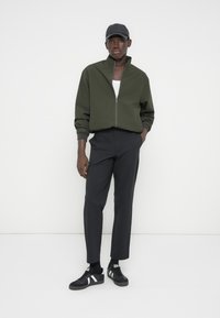 Νέος άνδρας που φοράει σκούρο πράσινο μπουφάν με φερμουάρ, μαύρο καπέλο, σκοτεινό παντελόνι και μαύρα αθλητικά παπούτσια με λευκές ρίγες στέκεται μπροστά σε λευκό φόντο.