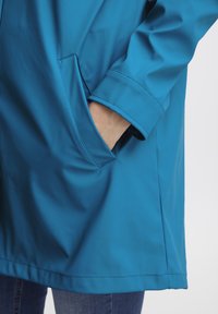 Blauwe waterdichte jas met een gladde textuur, zijzak en gestikte zoom. De mouw is uitgevoerd met een rechte snit.