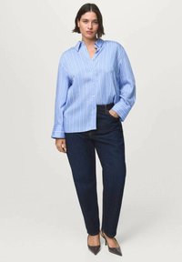 Camisa de botones a rayas azul claro con mangas largas, combinada con jeans de mezclilla oscura. La camisa tiene un corte holgado y un diseño clásico de cuello.
