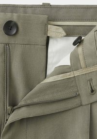 Gros plan de la ceinture d’un pantalon beige montrant un bouton, une fermeture éclair et une fermeture intérieure à crochet et barre partiellement ouverte sur un fond uni.