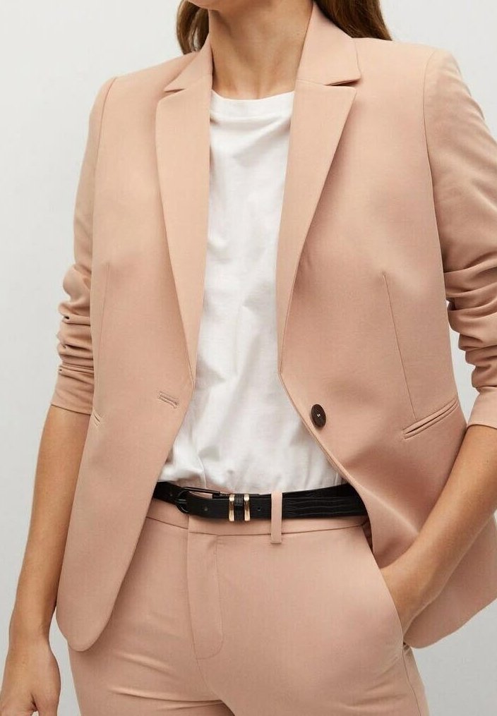 Femme portant un blazer beige clair ajusté et un pantalon assorti, avec une chemise blanche et une ceinture noire, les mains dans les poches.