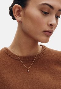Złoty naszyjnik z zawieszką w kształcie gwiazdy noszony na brązowym swetrze z dzianiny. Proste kolczyki typu sztyft uzupełniają stylizację. Czyste tło podkreśla biżuterię.
