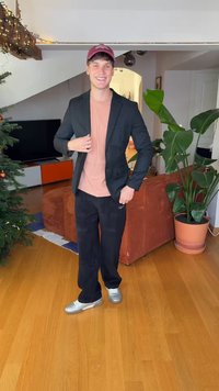 Blazer noir sur une chemise rose clair, assorti à un pantalon noir et des baskets argentées. Debout sur un sol en bois, avec des plantes et une décoration en arrière-plan.