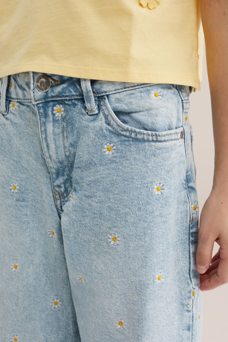 Jean en denim bleu clair avec de petites marguerites blanches et jaunes brodées, porté avec un haut jaune, main reposant sur le côté.
