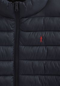 Granatowa kamizelka puchowa z zamkiem błyskawicznym, pikowaną fakturą i małym czerwonym haftowanym logo golfisty po lewej stronie.