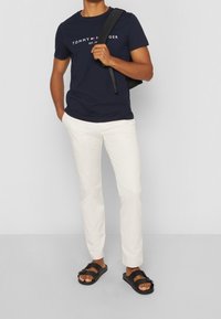 Camiseta de algodón azul marino con el texto blanco "TOMMY HILFIGER", combinada con pantalones ajustados blancos y sandalias de estilo slide negras.