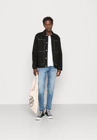 Chaqueta de mezclilla negra con costuras blancas, camiseta blanca, jeans azul claro y zapatillas negras altas. Lleva una bolsa beige con un diseño gráfico.