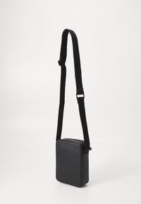 Černá crossbody taška obdélníkového tvaru, vyrobená z hladkého materiálu. Má dlouhý nastavitelný popruh a zipové uzavření.