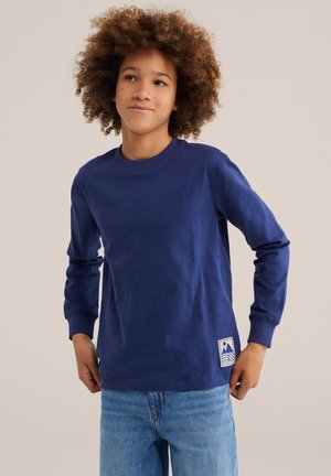 Jeune personne aux cheveux bouclés portant une chemise à manches longues bleue et un jean bleu clair sur un fond uni.