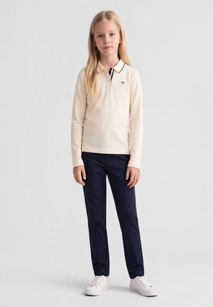 Fată tânără stând purtând un tricou polo bej cu mâneci lungi, pantaloni bleumarin și adidași albi pe un fundal simplu gri deschis.
