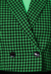 Vihreä ja musta houndstooth-kuvioitu bleiseri, jossa on korostetut liepeet, kaksoispainikkeet ja sileä pinta, kerroksittain vihreän paidan päällä.