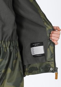Fleeceforet jakke i grøn camouflage med elastisk talje, lynlåslukning og indvendig labels til personlige oplysninger. Glat tekstur.
