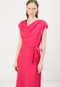 Rózsaszín, cowl nyakú ruha rövid ujjal, a deréknál megkötve, sima textúrával és lágy sziluettel, semleges háttér előtt.