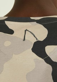 Camiseta de algodón con un patrón de camuflaje en beige, negro y gris, con un detalle de costura negra en el cuello trasero.