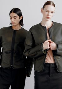 Zwei Modelle tragen dunkelgrüne Lederjacken mit abgerundeten Kanten. Die Jacken haben einen Reißverschluss und schwarze Akzente und sind mit minimalistischen Oberteilen kombiniert.