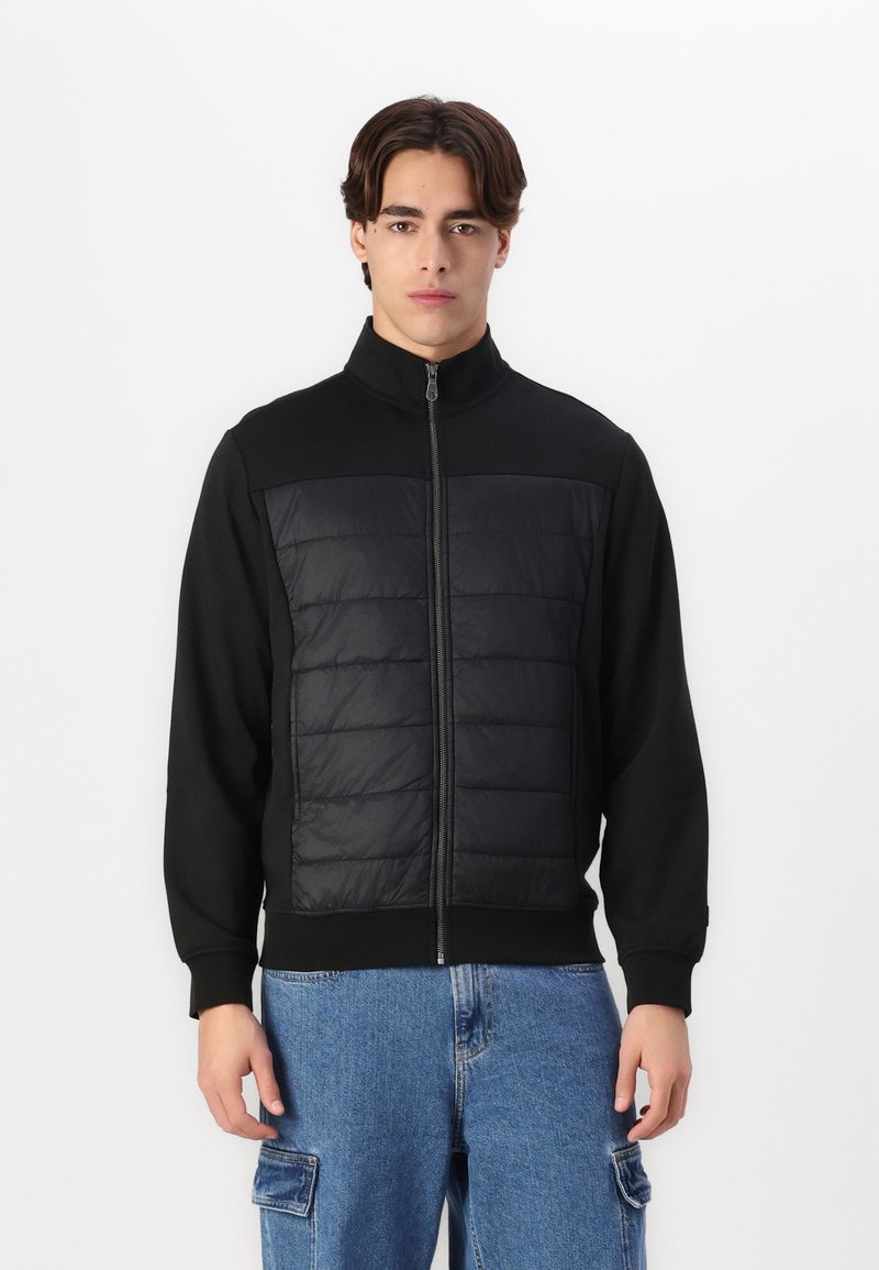 Svart jakke med høy krage, glidelås, med en polstret frontpanel. Glatt stoff med ribbestrikkede mansjetter og kant, båret over lyseblå jeans.