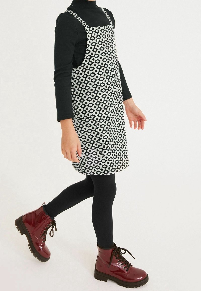 Enfant portant un haut à manches longues noir, des leggings noirs, une robe tablier fleurie noire et blanche, et des bottines à lacets rouge foncé brillant, en train de marcher.