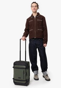 Brun mockaklädjacka med vit söm, mörkblå jeans, beige skor och en grön och svart väskvagn med dragkedjor.