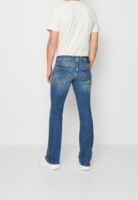 Jeans de denim azul con un corte recto y un ligero desgaste. Llevados con una camisa de manga corta de color claro y con puntos, y zapatillas blancas.