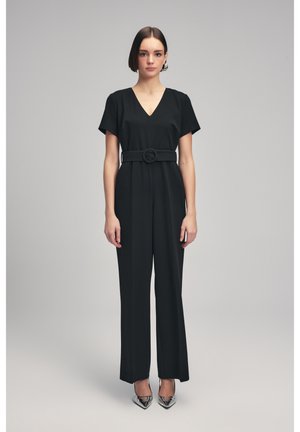 Jumpsuit noir avec un décolleté en V, des manches courtes, une taille ceinturée avec une boucle ronde, et un pantalon large ; modèle portant des talons argentés.