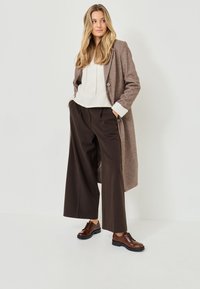 Abrigo de lana marrón con diseño de doble botonadura, combinado con una sudadera blanca con capucha y pantalones anchos marrones. Unos zapatos de cordones marrones completan el atuendo.