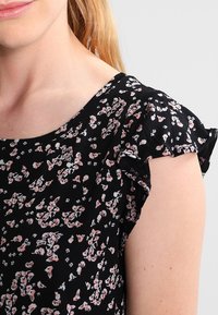 Svart blommig topp med små rosa och vita blommor, korta volangärmar och rundad halsringning. Lättviktig tyg och strukturerad mönster.