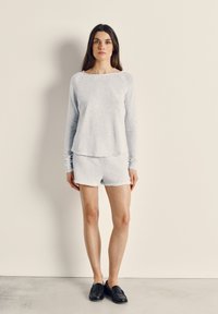 Pikkade tumedate juustega naine, kes kannab heledat halli pika varrukaga toppi ja sobivaid lühikesi pükse, seisab üksluisel beežil seinapinnal, jalas tal mustad mokassiinid.