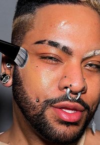 Pennello per trucco che applica il prodotto sulla pelle con una lucentezza visibile. In primo piano, viso con barba scura, molteplici piercing e pelle con texture.